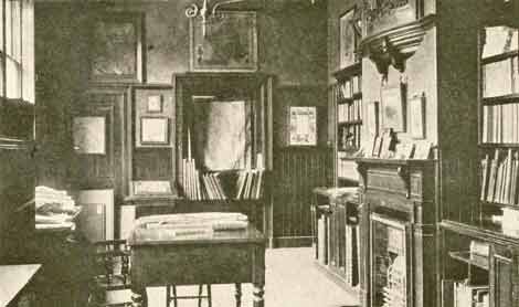 Early photo of a room with bound books on shelves, fireplace, table and chair